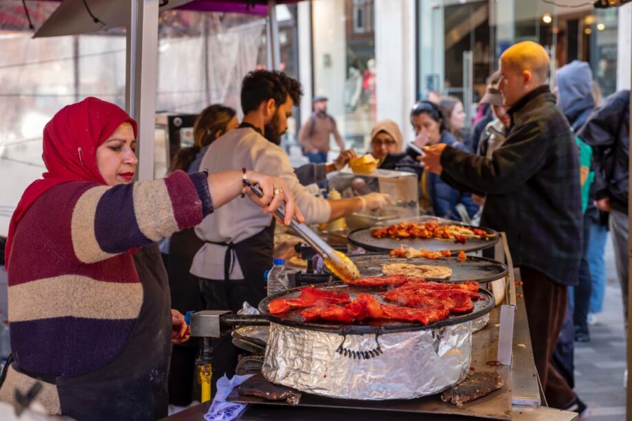 Seasoned chicken on large circle grill being cooked by man in a branded apron.
