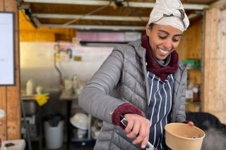 Ethiopian lady handing a pot of food to customer in a brown cardboard container.